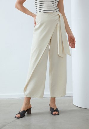 Vrouw draagt crèmewitte wijde broek met asymmetrisch overslagrokdetail en zwarte open teen sandalen met hak op een betonnen vloer.