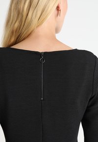 Svart texturerad topp med horisontell ribbing, en bred halsringning och en synlig central dragkedja bak för stängning.