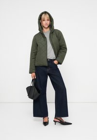 Giacca a vento quilted verde con cappuccio, camicia grigia, jeans in denim scuro a gamba larga, tacchi slingback neri e borsa nera. Design minimalista con linee pulite.