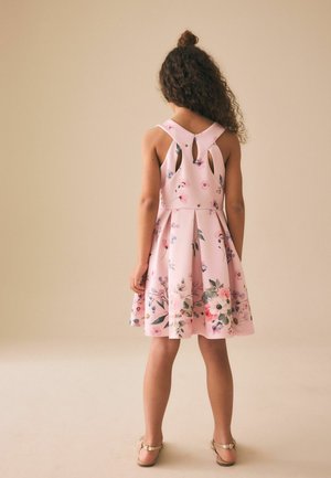 Neiu lokkis juustega, juuksed seotud sõlme, seljas roosad lillemustriga varrukateta kleit, millel on seljal lõiked, ja kuldsed sandaalid, seisab beežil põrandal.