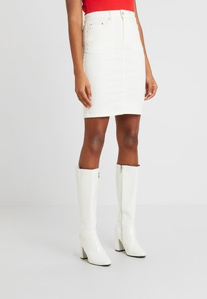 Frau trägt einen weißen Jeansrock, kniehohe weiße Stiefel mit Absatz und ein rotes ärmelloses Oberteil vor einem einfarbigen Hintergrund.