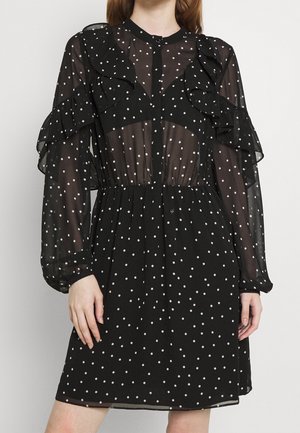 Robe noire transparente à pois blancs, avec des volants sur les épaules, des manches longues et une taille élastique, portée par une personne debout.