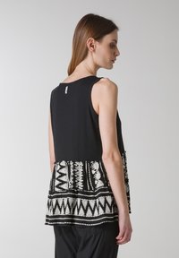 Sort tanktop med et mønstret peplum-hem med hvide tribal motiver. Bagsiden har en rund udskæring og et synligt mærke.