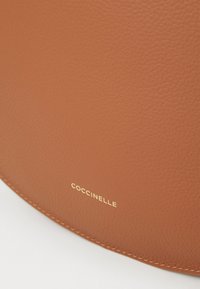 Kohvipruun nahast kott, millel on tekstureeritud pind, ümar kuju ja kuldne "Coccinelle" reljeefne logo allosas.