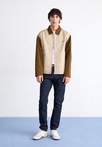 Beige en bruine zip-up jas met een kraag, met lange mouwen. Draagt over een wit shirt en gecombineerd met donkerblauwe jeans en crème sneakers.