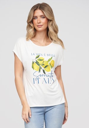 Kvinde med blondt hår iført en hvid t-shirt med citroner og teksten ""La Vita È Bella Sorrento Italy"" og lyseblå jeans.