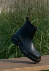 Μαύρη δερμάτινη μποτάκι Chelsea με ελαστικές πλαϊνές επενδύσεις, στρογγυλή μύτη και χαμηλό τετράγωνο τακούνι, κρεμασμένο πάνω από μια ξύλινη επιφάνεια, σε φόντο με πρασινάδα.