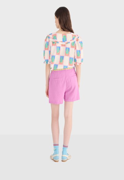 Femme portant un chemisier à motifs avec un col à volants, un short rose taille haute, des chaussettes bleues et des chaussures crème, debout de dos.