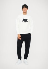 Hvit hettegenser med svart logo, kombinert med svarte joggebukser og hvite joggesko. Enkel, uformell design uten mønstre eller ekstra detaljer.