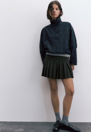 Jeune femme portant un coupe-vent foncé, une jupe plissée, des chaussettes rayées et des mocassins noirs, se tenant contre un mur blanc uni.