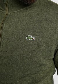 Zelený svetr na zip vyrobený z texturované tkaniny, s malým vyšitým logem krokodýla na hrudi.