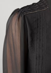 Manche de blouse noire en tissu transparent, plis verticaux et petite bordure de perles argentées le long de la couture de l'épaule.