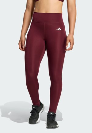 Mujer con leggings burdeos de talle alto y zapatillas negras Adidas, de pie con las piernas ligeramente separadas sobre un fondo claro.