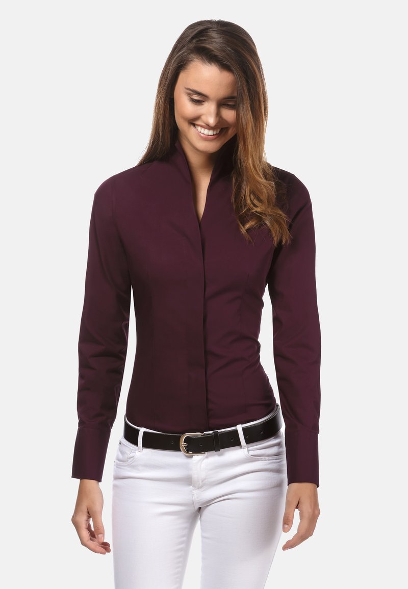 Bordeauxrood shirt met lange mouwen, hoge kraag en een gladde textuur, in combinatie met witte broek en een zwarte riem.