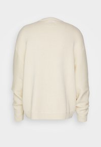 Pull crème en maille texturée, avec des épaules tombantes et un ourlet côtelé. Il présente un col rond et une coupe décontractée. Pas de fermetures visibles.