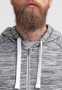 Persona con barba e tatuaggi sul collo che indossa una felpa grigia con zip e lacci bianchi su uno sfondo semplice.
