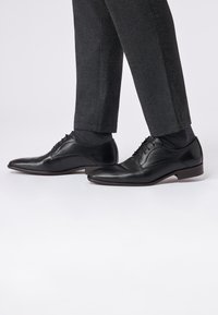 Schwarze Lederdressschuhe mit glatter Oberfläche, ausgestattet mit dekorativen Perforationen und Schnürsenkeln. Kombiniert mit dunklen maßgeschneiderten Hosen.