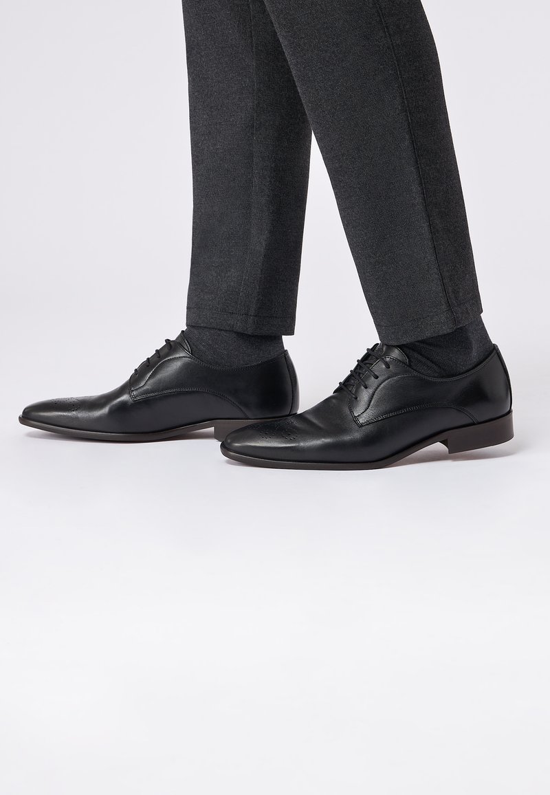 Schwarze Lederdressschuhe mit glatter Oberfläche, ausgestattet mit dekorativen Perforationen und Schnürsenkeln. Kombiniert mit dunklen maßgeschneiderten Hosen.