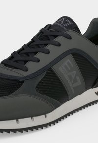 Baskets gris foncé et noires avec panneaux texturés, fermeture à lacets, semelle blanche rainurée et marquage "EA7 Emporio Armani" sur le côté et la languette.
