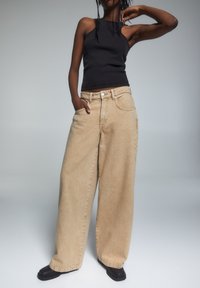 Beżowe spodnie dżinsowe z szerokimi nogawkami i wysokim stanem, z przednimi kieszeniami i subtelnym przeszyciem, połączone z dopasowanym czarnym topem na ramiączkach.