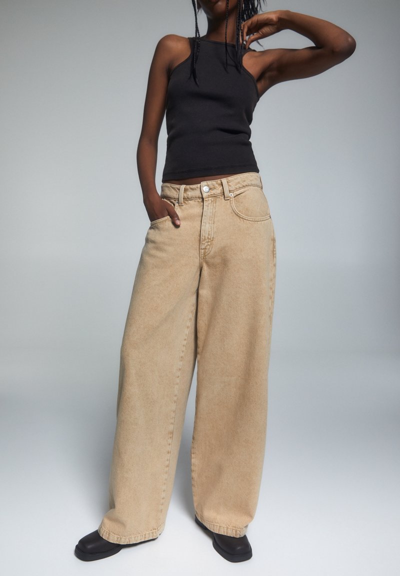 Beżowe spodnie dżinsowe z szerokimi nogawkami i wysokim stanem, z przednimi kieszeniami i subtelnym przeszyciem, połączone z dopasowanym czarnym topem na ramiączkach.