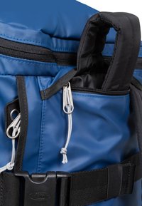 Sininen kangaskassi, jossa on musta vetoketju, säädettävät hihnat ja kestävä solki. Sisältää silmukalla varustetun kahvan ja useita kiinnityspisteitä.