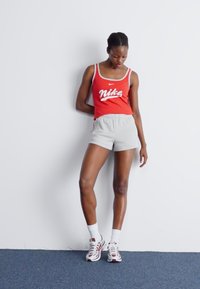 Raudonas Nike marškinėlių topas su baltu logotipu, pilkos šortai, balti kojinės ir pilkai-raudoni sportbačiai. Modelis stovi prieš baltą sieną.