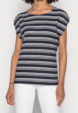 Vrouw draagt een donkerblauw-wit horizontaal gestreept top met korte mouwen en knoopdetails op de schouders, gecombineerd met een witte broek, tegen een effen achtergrond.