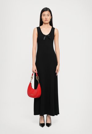 Woman in a sleeveless black maxi dress with a small bow, holding a red handbag with gold chain handles, wearing black pointed heels.