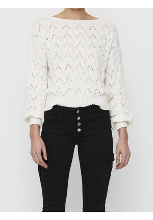 Vrouw draagt een wit gebreide trui met een chevronpatroon en zwarte hoge taillebroek met vier zichtbare knopen en zijzakken.