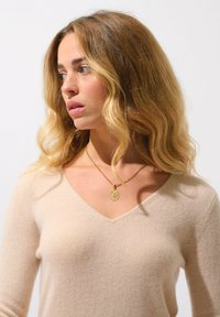 Krémový svetr s výstřihem do V vyrobený z měkké látky, s jemnou strukturou. Zlatý náhrdelník s přívěskem s ozdobným designem, nošený kolem krku.