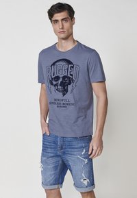 Pilkos medvilninės marškinėliai su kaukolės grafika ir užrašu "RUGGED". Derinama su mėlynomis džinsinėmis šortais, turinčiomis pablogintus išsiskyrimus ir surištus kraštus.