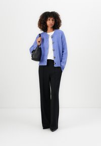 Šviesiai violetinė megzta kardiganas su apvaliu iškirpimu ir sagų uždarymu, derinamas su baltu marškinėliu ir juodomis plačiomis kelnėmis. Juoda pečių rankinė.