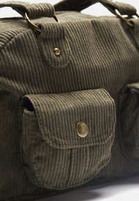 Khaki kordfløjl taske med to frontlommer, guldfarvede detaljer og ribbet tekstur. Har runde håndtag og lynlås lukning.