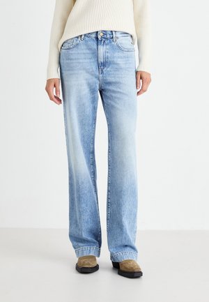 Osoba ubrana w jasnoniebieskie szerokie spodnie dżinsowe, kremowy prążkowany sweter oraz brązowe buty na kwadratowym nosku stoi na białej podłodze.