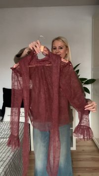 Blusa di pizzo color bordeaux con tessuto trasparente, maniche arricciate e fiocco al collo. Esibita su una gruccia contro uno sfondo chiaro.