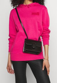 Felpa rosa brillante con tasca frontale, indossata con pantaloni neri in ecopelle. Una piccola borsa a tracolla nera ha una chiusura a pattina e un logo.