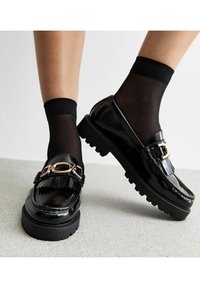 Mocasines de charol negro con herrajes dorados, detalle de correa y una suela gruesa y texturizada; combinados con calcetines negros transparentes.