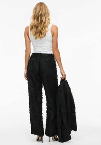 Black textured pants with a floral design; paired with a matching jacket held in hand. White sleeveless top and heels complete the look.