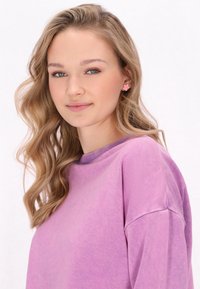 Felpa a maniche lunghe, di colore viola, con scollo rotondo e una texture morbida. Indossata con piccoli orecchini a forma di fiore rosa. Sfondo chiaro.