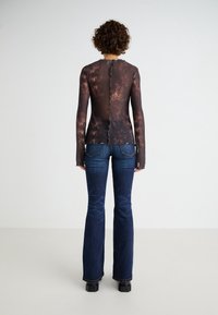 Blusa transparente de manga larga con un patrón de tie-dye marrón, bordes en forma de concha y cierre de botón en la parte posterior, combinada con unos pantalones vaqueros acampanados de color azul oscuro.