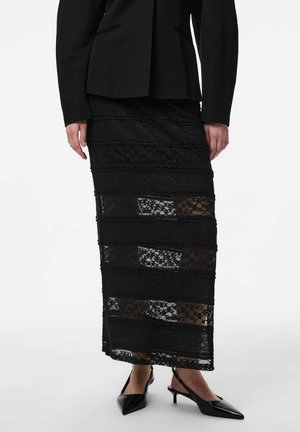 Femme portant une jupe midi en dentelle noire avec des panneaux transparents horizontaux, un blazer noir ajusté, et des chaussures slingback noires à talons bas et bouts pointus.