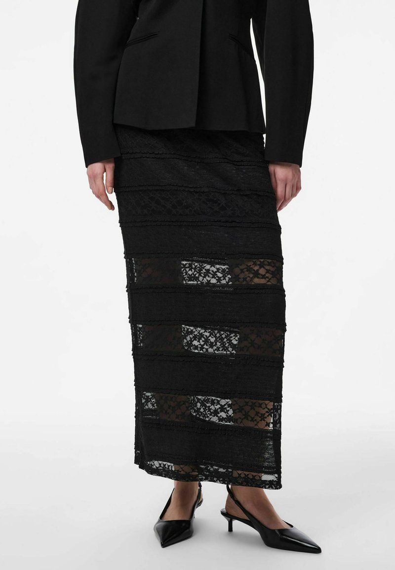 Femme portant une jupe midi en dentelle noire avec des panneaux transparents horizontaux, un blazer noir ajusté, et des chaussures slingback noires à talons bas et bouts pointus.