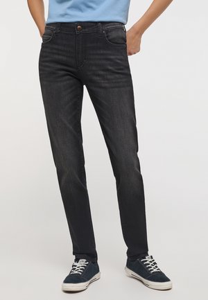 Homme portant un jean noir ajusté et des baskets bleu marine avec des lacets blancs, debout avec une main dans la poche arrière sur un fond uni.