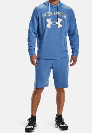 Felpa blu in misto cotone con logo "Under Armour" bianco, abbinata a pantaloni corti blu coordinati. Scarpe da ginnastica nere con dettagli rossi.