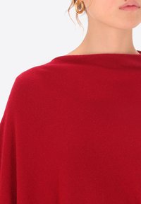 Donna che indossa grandi orecchini a cerchio d'oro e un top lavorato a maglia rosso con scollatura ampia, vista dalla mascella alla spalla.