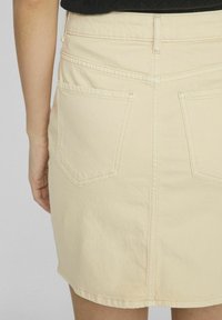 Falda de mezclilla beige con una cintura ajustada, dos bolsillos traseros, costuras visibles y un dobladillo recto. El material parece ligeramente texturizado.