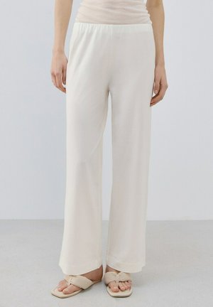 Mujer con pantalones anchos color crema y sandalias anudadas a juego, de pie contra un fondo liso y claro.