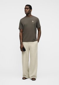 Άνδρας που στέκεται, φορώντας ελιάς πράσινο T-shirt, μπεζ παντελόνι με φαρδιά πόδια, μαύρα σανδάλια, κρατώντας ένα μικρό μαύρο πορτοφόλι στο δεξί χέρι.