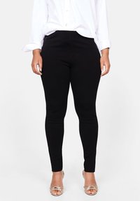 Svarta leggings i en åtsittande design, tillverkade av elastiskt tyg, i kombination med en vit skjorta med knappar och ljusa sandaler.